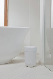 Weißer Mülleimer mit Deckel, im Badezimmer neben einer Badewanne platziert, aus seitlicher Perspektive fotografiert.