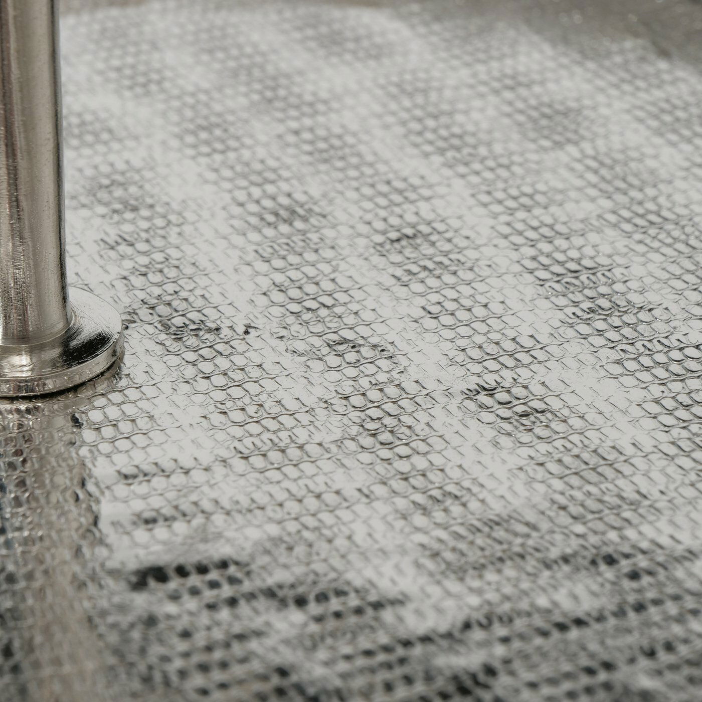 Nahaufnahme einer strukturierten, silbernen Oberfläche mit einem Metallstab, perspektivisch von der Seite betrachtet.