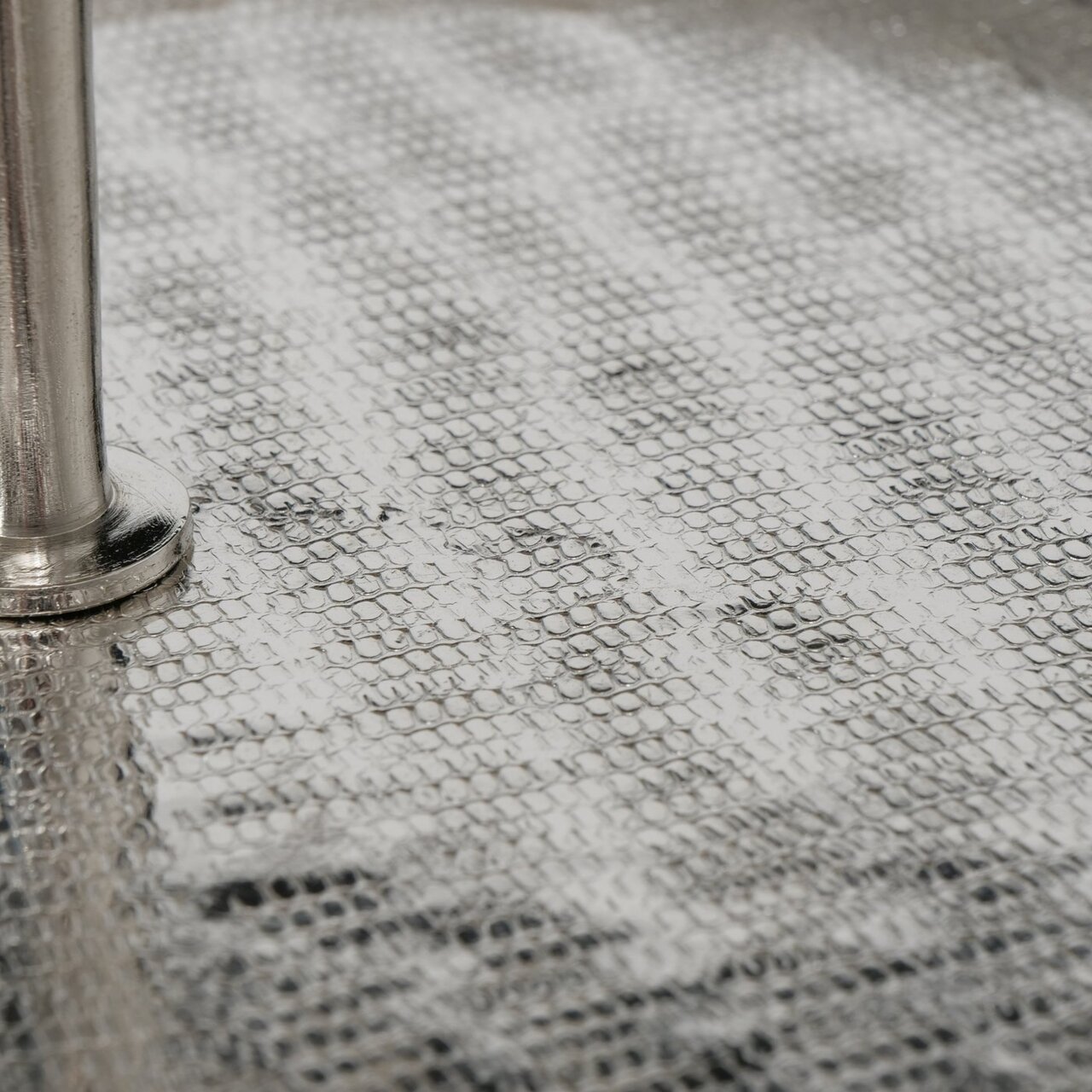 Nahaufnahme einer strukturierten, silbernen Oberfläche mit einem Metallstab, perspektivisch von der Seite betrachtet.