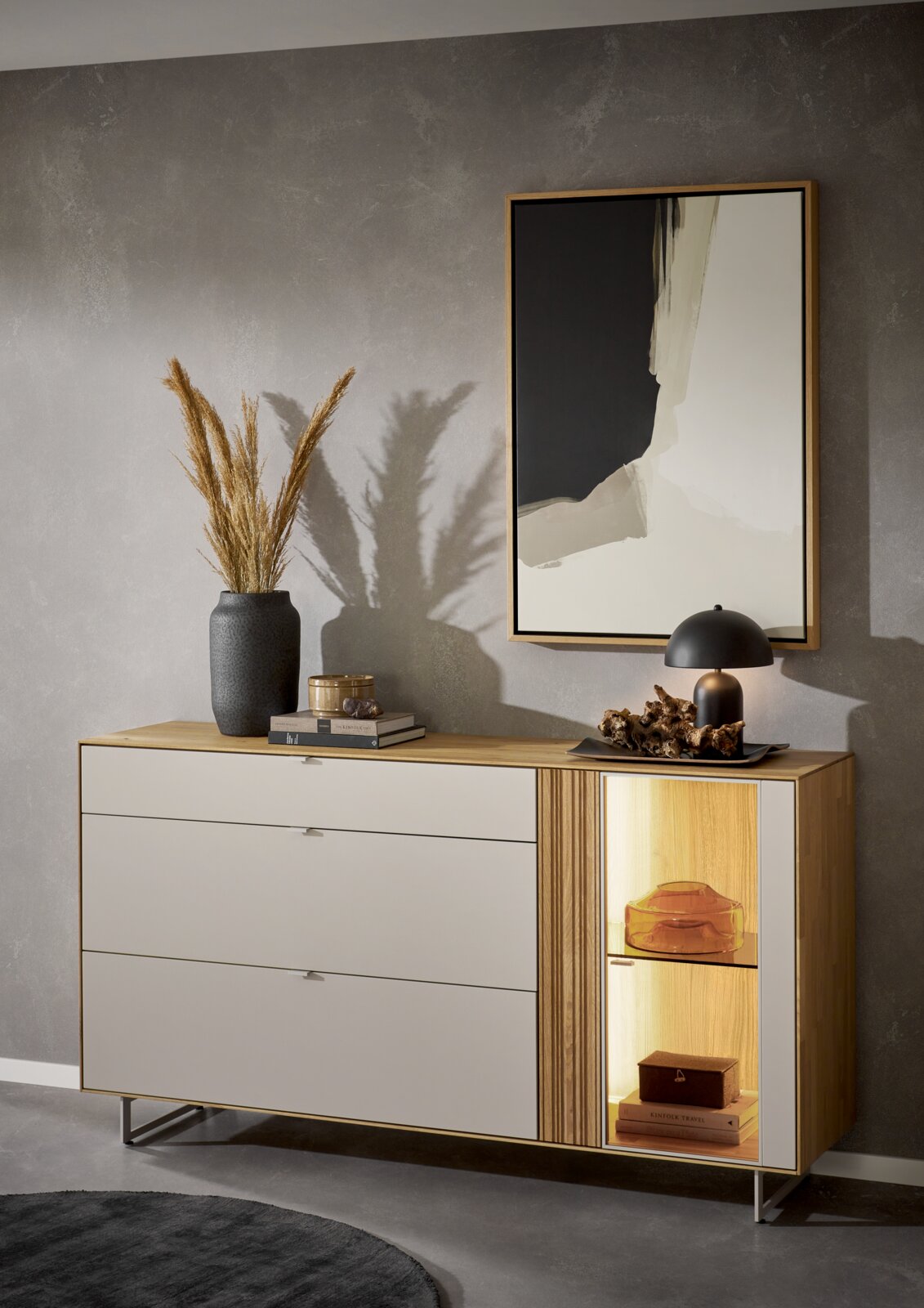 Moderne Sideboard-Kommode in Weiß mit Holzoberfläche und beleuchtetem Glasvitrinenfach rechts, dekoriert mit Vase, Büchern und Tischlampe; Perspektive: frontale Ansicht mit leichter Schrägsicht von links.