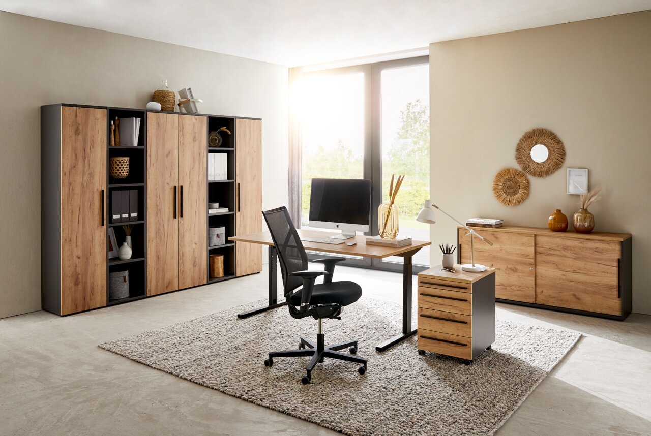 Modernes Büro mit Holzmöbeln, bestehend aus einem Schreibtisch, einem Rollcontainer und mehreren Aktenschränken. Die Perspektive zeigt den Raum von vorne mit Blick auf das Fenster im Hintergrund.