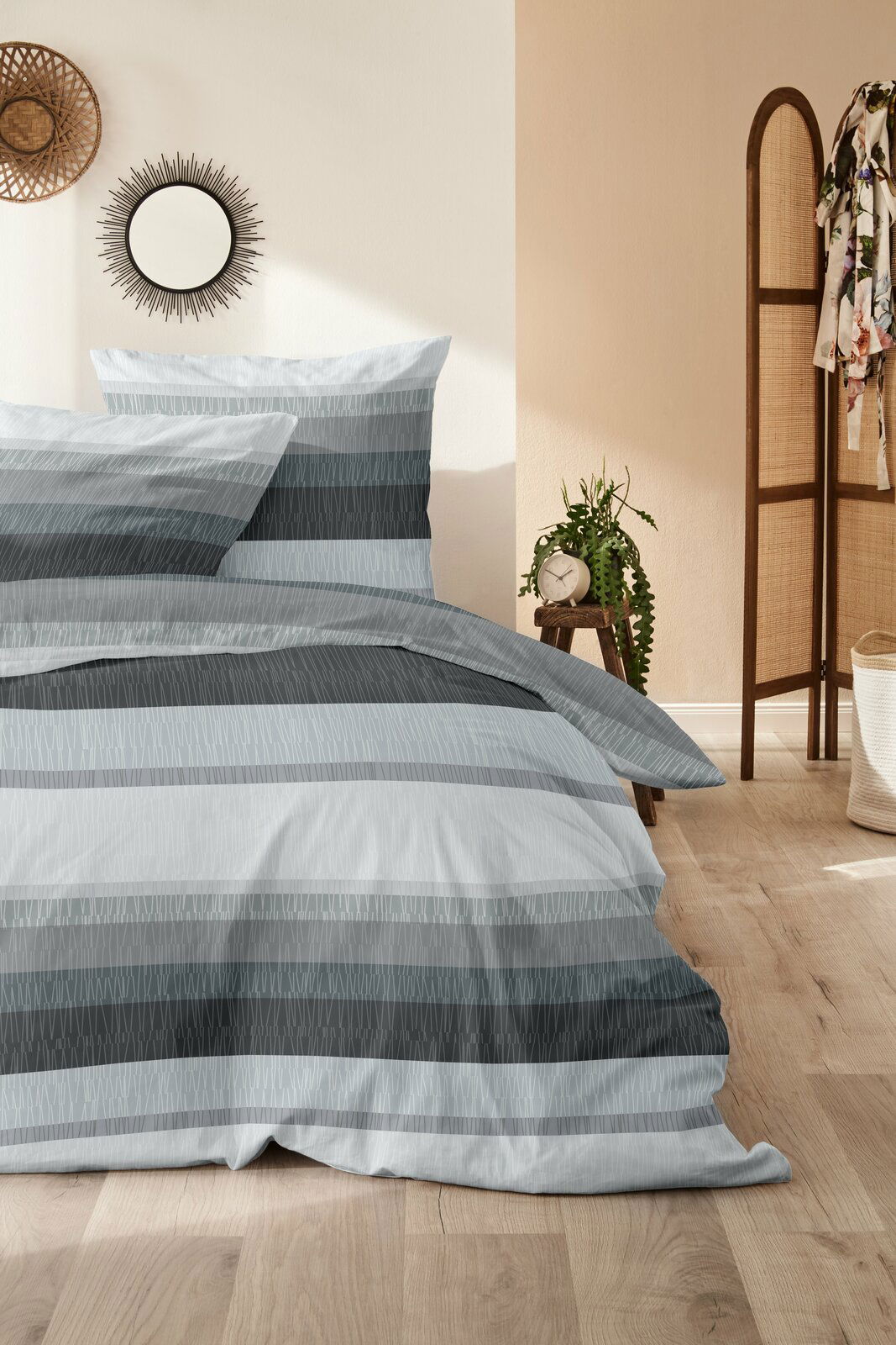 Bettwäsche-Set in Grau- und Blautönen mit Streifenmuster, auf einem Bett in einem stilvoll eingerichteten Raum mit Holzboden und Dekorationen, aus der Perspektive von vorne fotografiert.