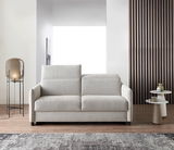 Modernes Schlafsofa in hellem Beige, frontal fotografiert, mit eleganten Armlehnen und einem gemütlichen Design, platziert in einem stilvollen Wohnzimmer mit Lampe und Beistelltischen.
