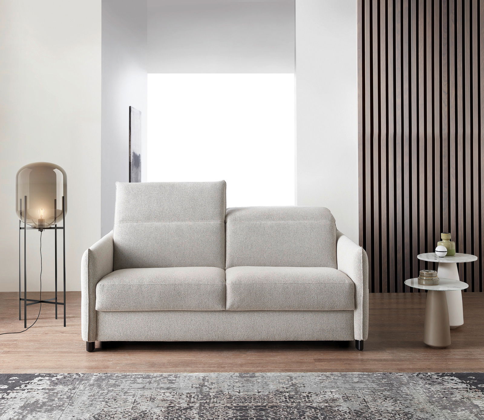 Modernes Schlafsofa in hellem Beige, frontal fotografiert, mit eleganten Armlehnen und einem gemütlichen Design, platziert in einem stilvollen Wohnzimmer mit Lampe und Beistelltischen.