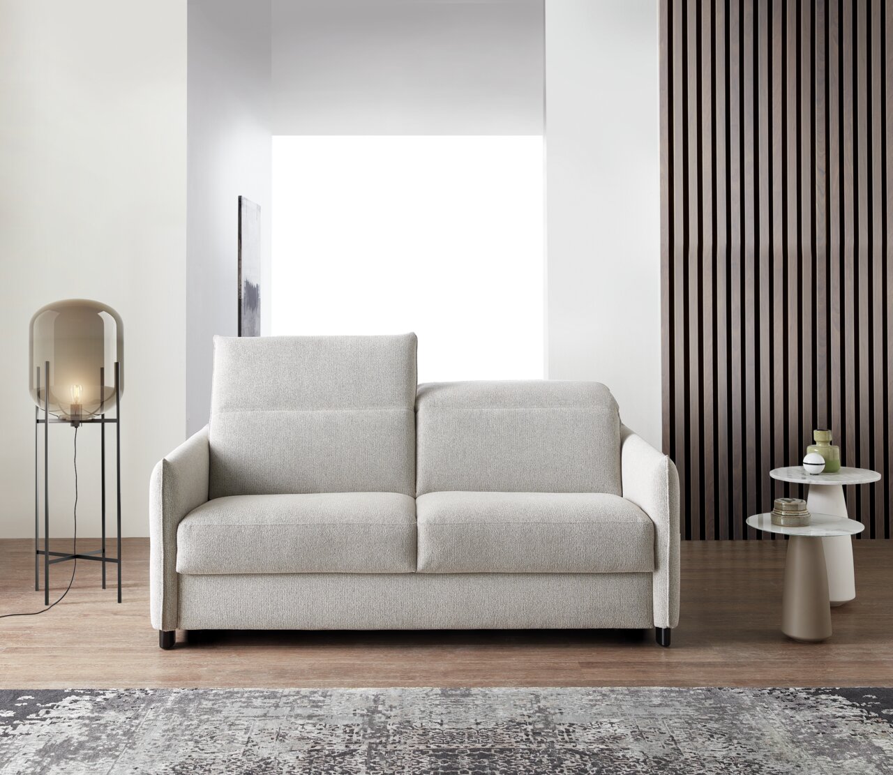 Modernes Schlafsofa in hellem Beige, frontal fotografiert, mit eleganten Armlehnen und einem gemütlichen Design, platziert in einem stilvollen Wohnzimmer mit Lampe und Beistelltischen.