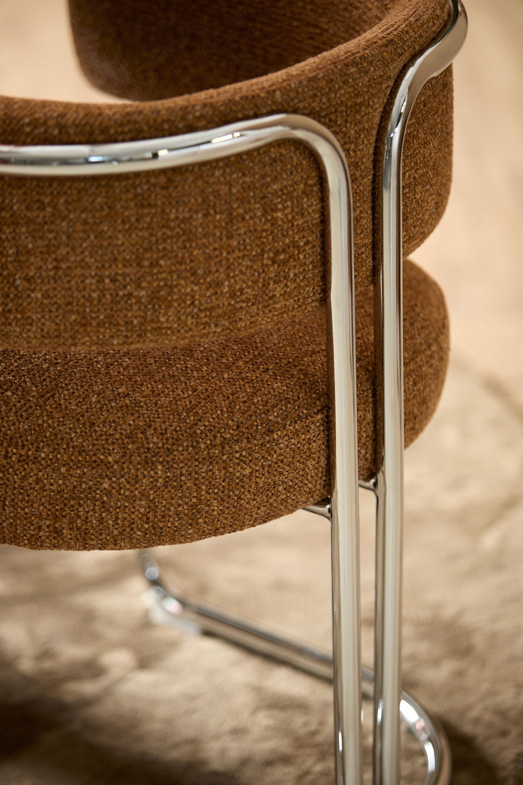 Nahaufnahme eines Armlehnstuhls mit braunem Stoffbezug und verchromtem Metallgestell, von hinten betrachtet.