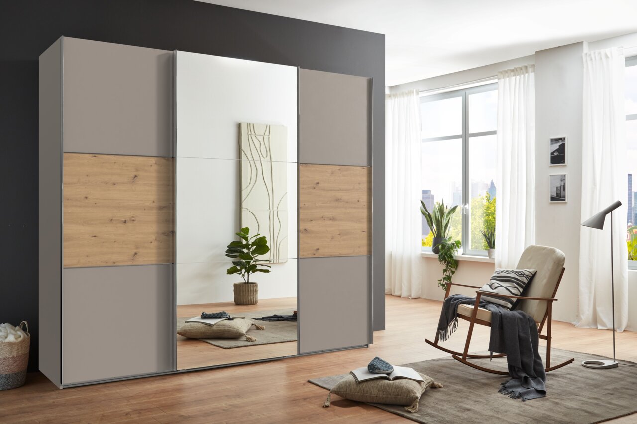 Moderner dreitüriger Schwebetürenschrank mit Holz- und Spiegelelementen, seitliche Perspektive im hellen Raum mit großen Fenstern.