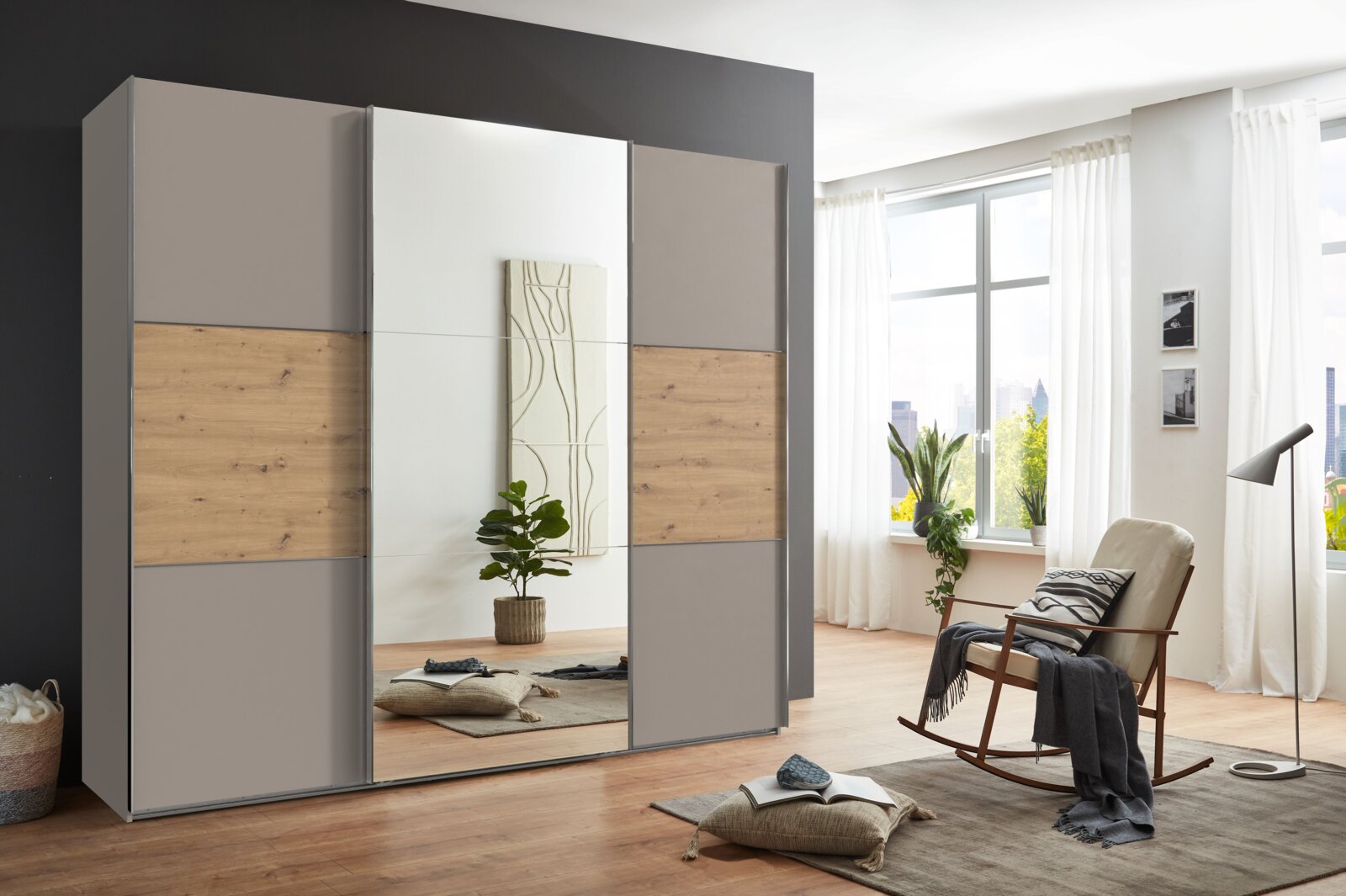 Z2 Schwebetürenschrank 3-trg.INDIANA A Moderner dreitüriger Schwebetürenschrank mit Holz- und Spiegelelementen, seitliche Perspektive im hellen Raum mit großen Fenstern.
