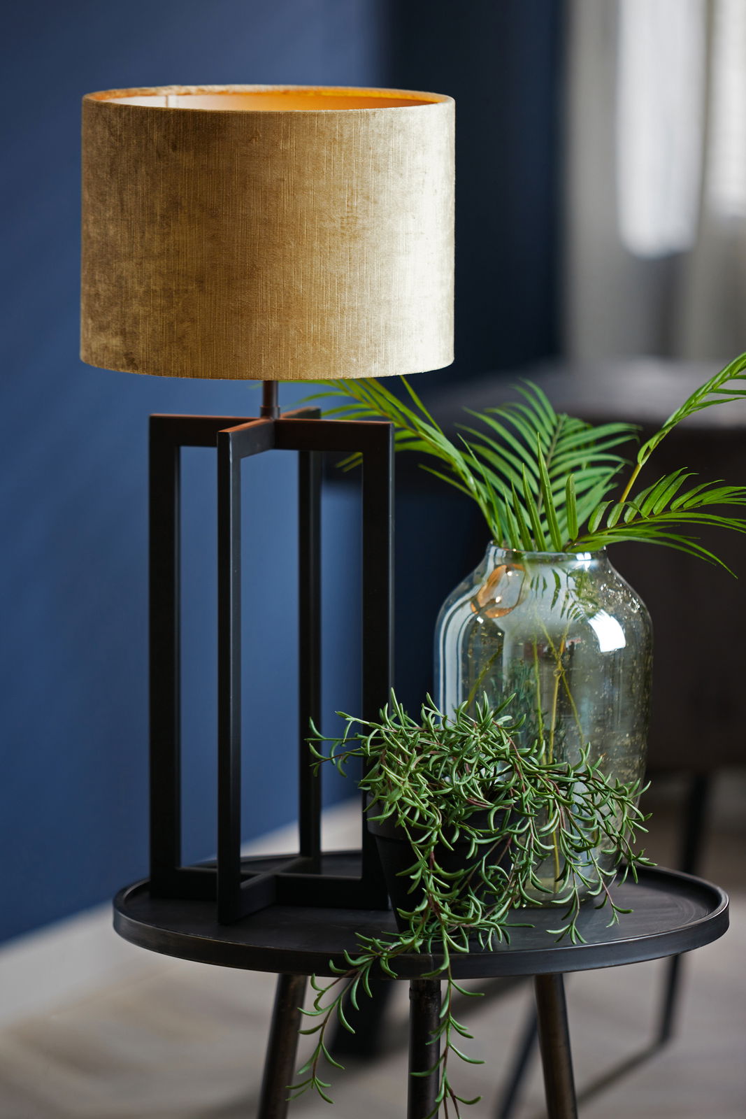 Moderner Lampenschirm in Zylinderform, goldfarben, auf einem schwarzen geometrischen Lampenfuß, umgeben von grünen Pflanzen, seitliche Perspektive.