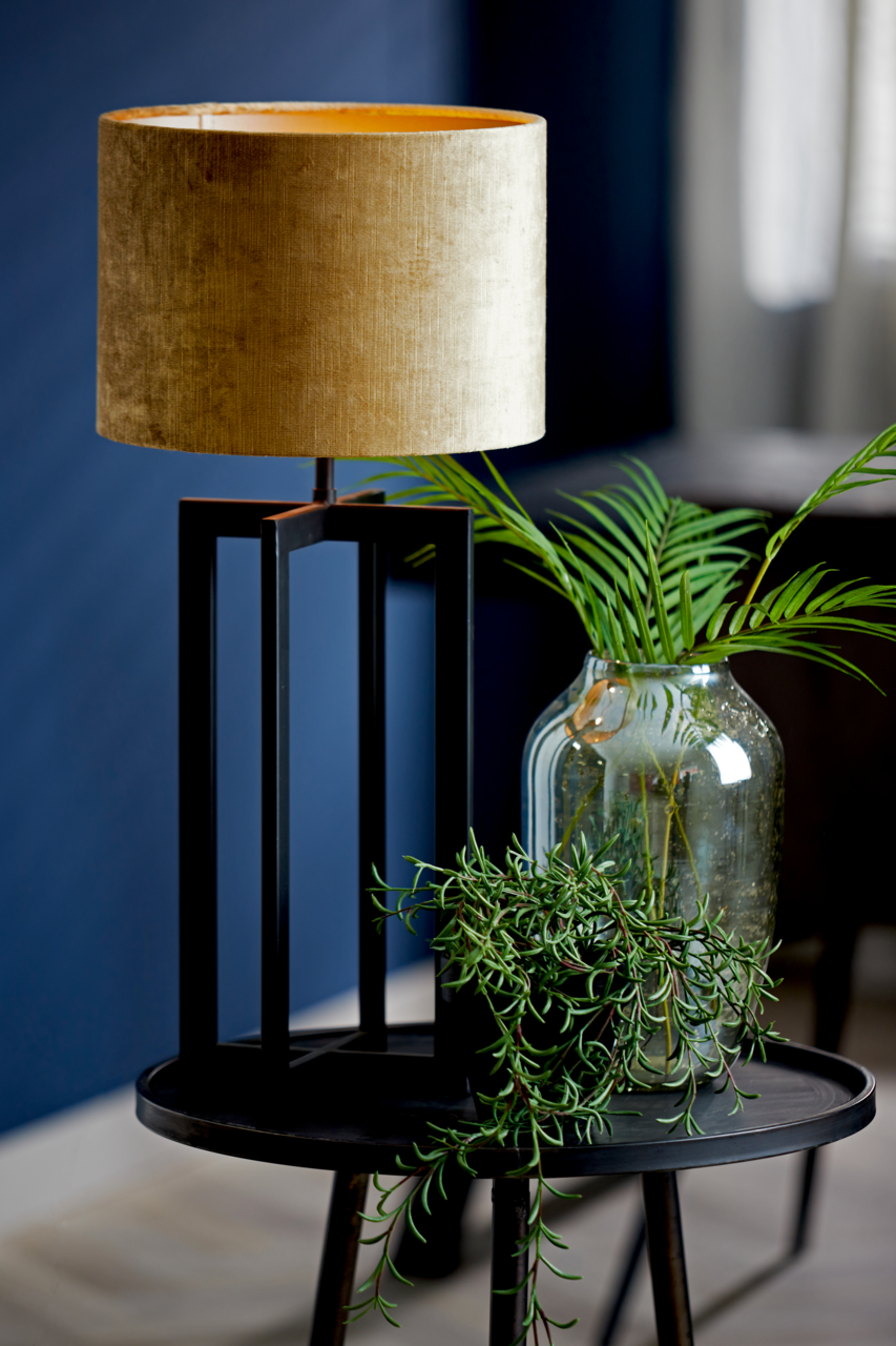 Moderner Lampenschirm in Zylinderform, goldfarben, auf einem schwarzen geometrischen Lampenfuß, umgeben von grünen Pflanzen, seitliche Perspektive.