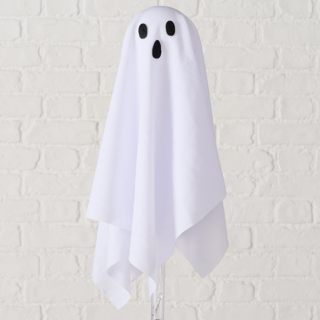 Weißes Halloween-Geister-Dekoelement mit schwarzem Augen- und Mundmotiv, frontal aus der Vorderansicht fotografiert vor heller Ziegelwand