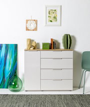 Frontale Innenraumaufnahme: weißes Sideboard mit Holzplatte und Schubladen, dekoriert mit Büchern, grüner Box und Kaktus; darüber zwei gerahmte Wandbilder an weißer Wand.