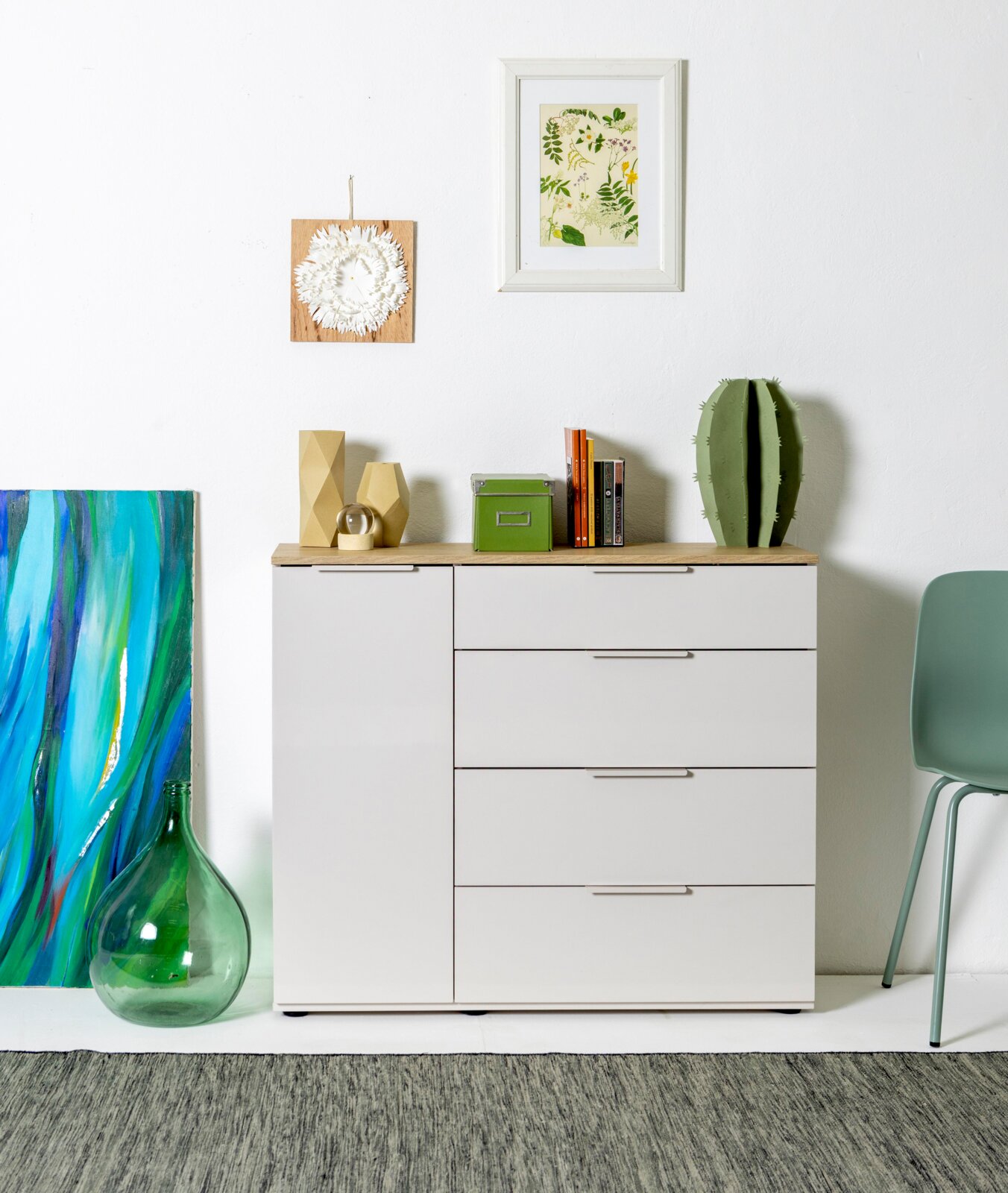 XORA Kommode MACCHIATO Frontale Innenraumaufnahme: weißes Sideboard mit Holzplatte und Schubladen, dekoriert mit Büchern, grüner Box und Kaktus; darüber zwei gerahmte Wandbilder an weißer Wand.