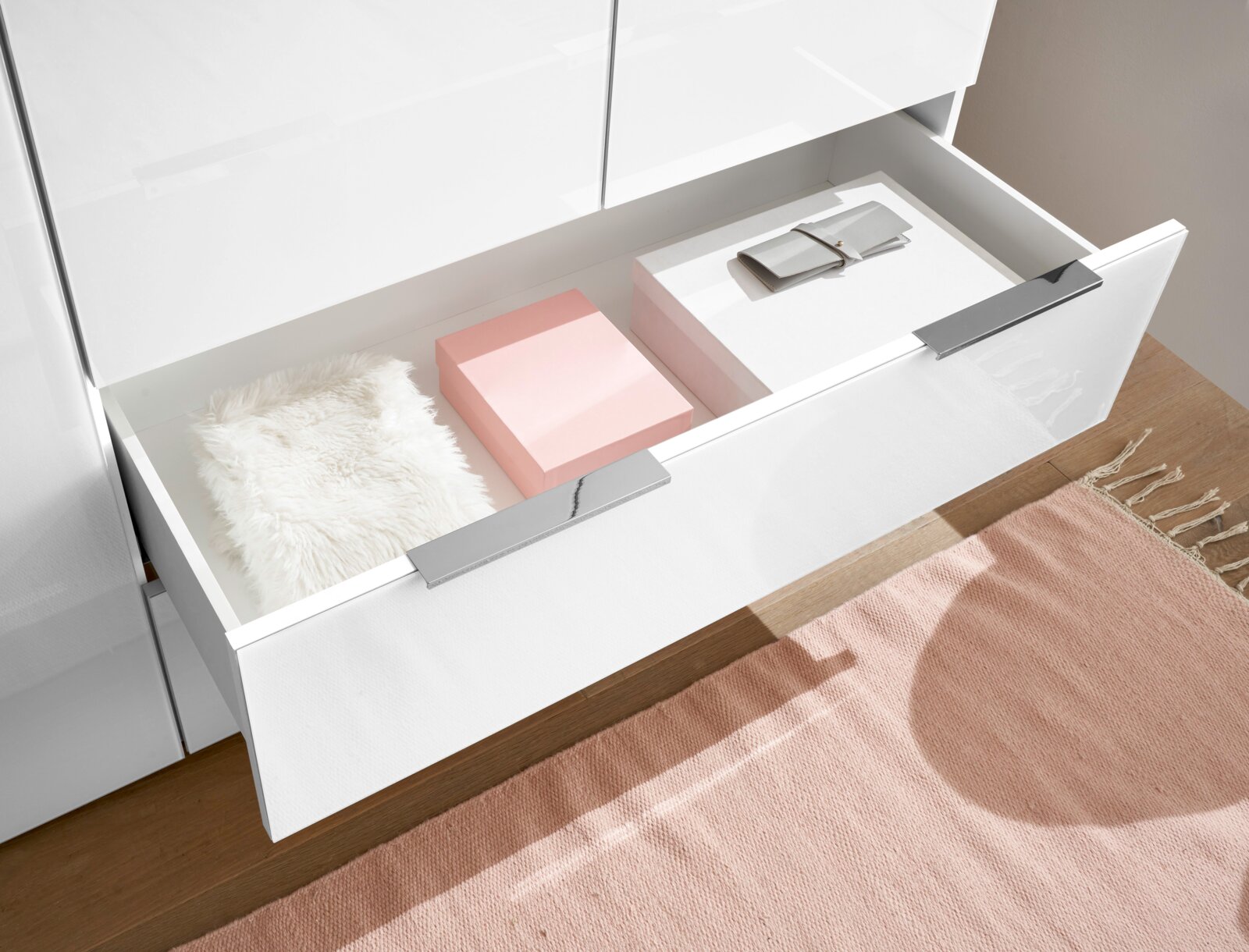 Geöffnete weiße Schublade eines modernen Schranks mit glänzenden Metallgriffen, innen mit rosa und weißer Box sowie kleinem Etui, perspektivisch in schräger Draufsicht von oben