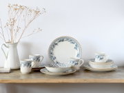Weißes Keramik-Geschirrset mit blauem floralen Muster (Teller, Schalen und Tassen) auf Holzregal vor weißer Wand, Frontansicht auf Augenhöhe.