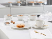 Helles, modernes Tischgedeck mit weißer Kaffeetasse auf Untertasse und goldenem Löffel, zwei Wassergläsern und Gebäck auf weißem Teller; Perspektive: leicht erhöhte Frontansicht (schräg von vorn).