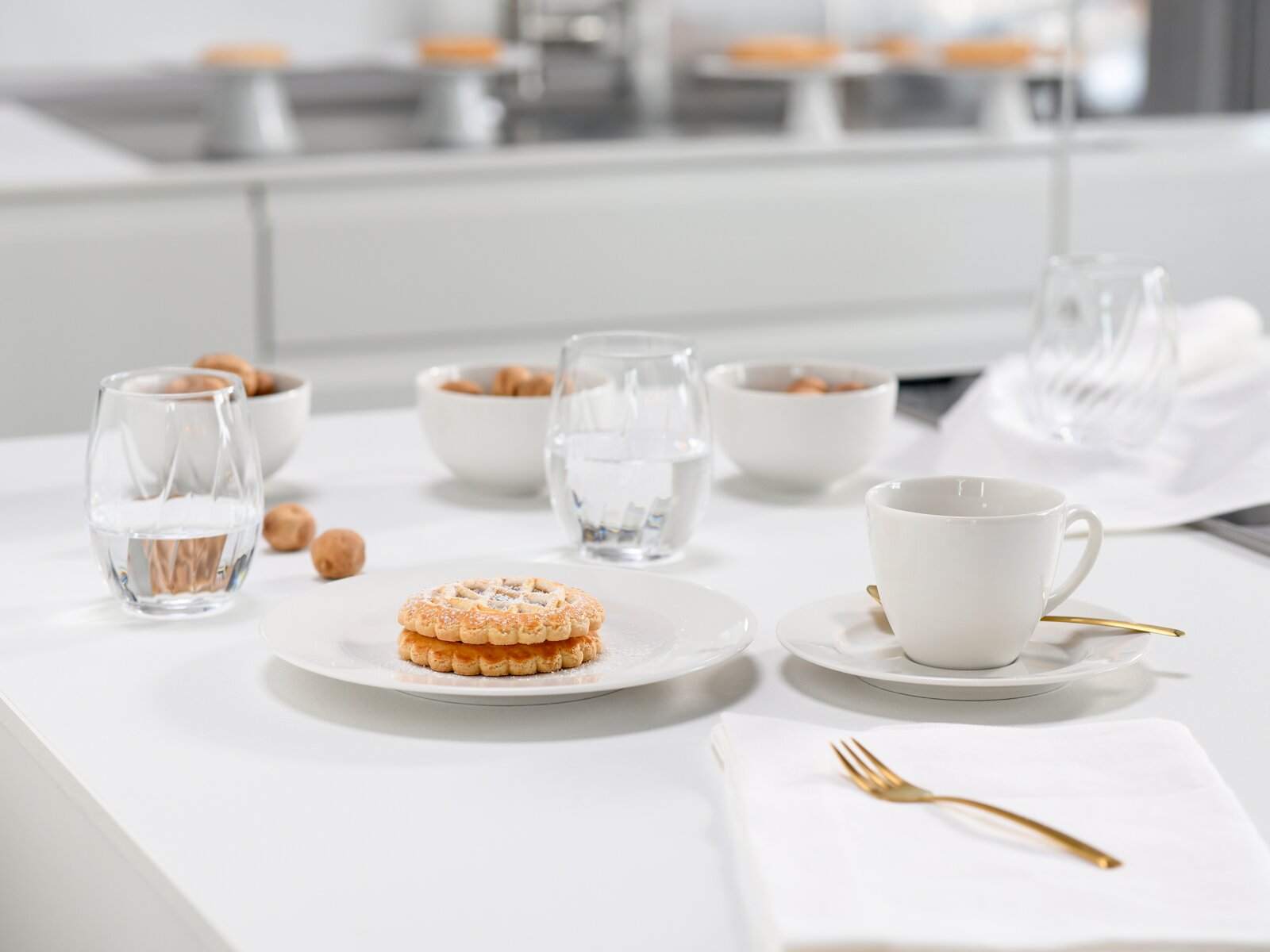 Helles, modernes Tischgedeck mit weißer Kaffeetasse auf Untertasse und goldenem Löffel, zwei Wassergläsern und Gebäck auf weißem Teller; Perspektive: leicht erhöhte Frontansicht (schräg von vorn).