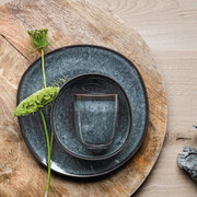Vogelperspektive auf einen grauen Speiseteller Lave 28cm mit passendem Becher und Schale, dekoriert mit einer grünen Blume auf einem Holztisch.
