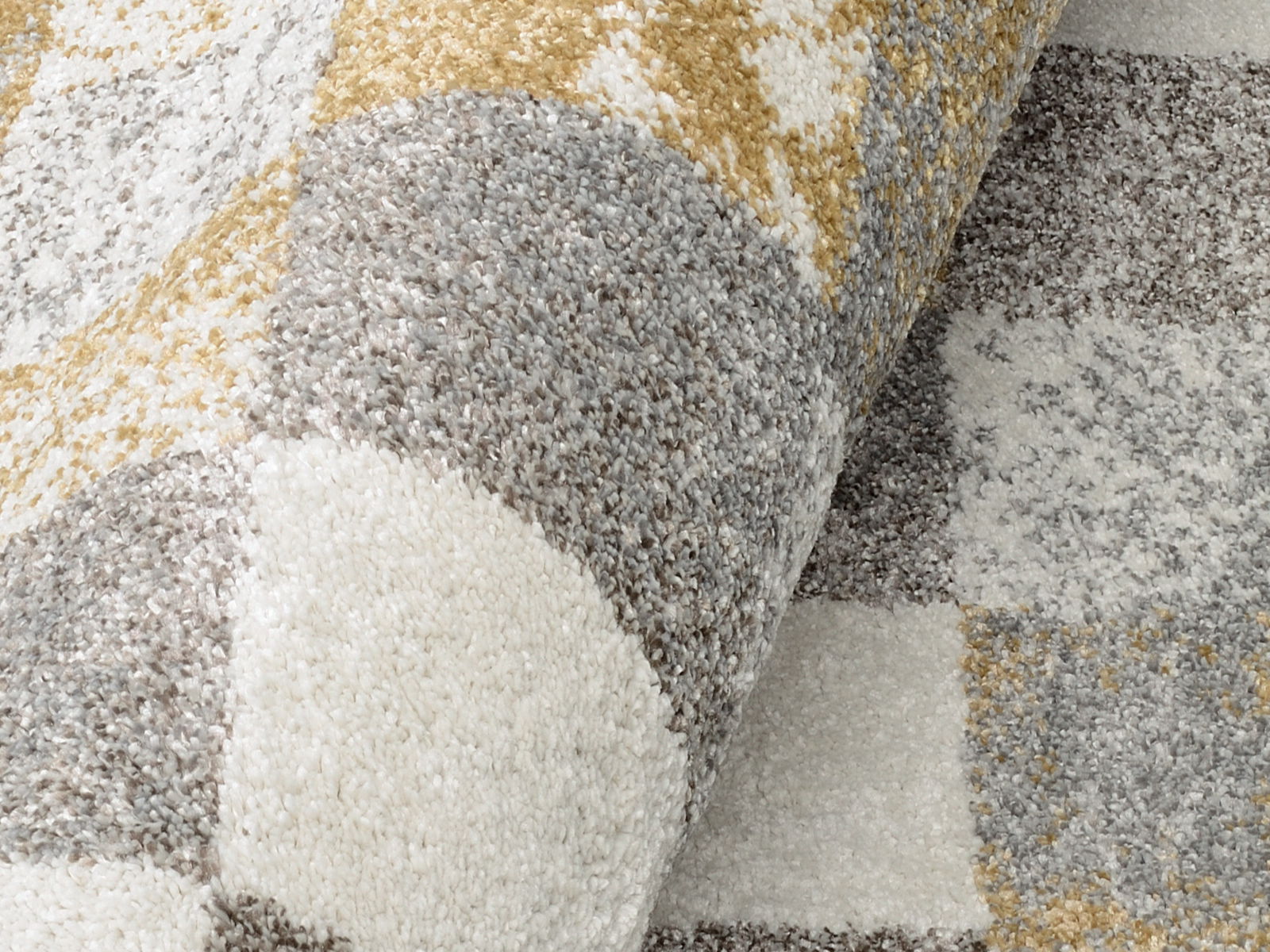 Nahaufnahme eines Frisé-Teppichs mit geometrischem Muster in Grau-, Weiß- und Goldtönen, leicht aufgerollt.