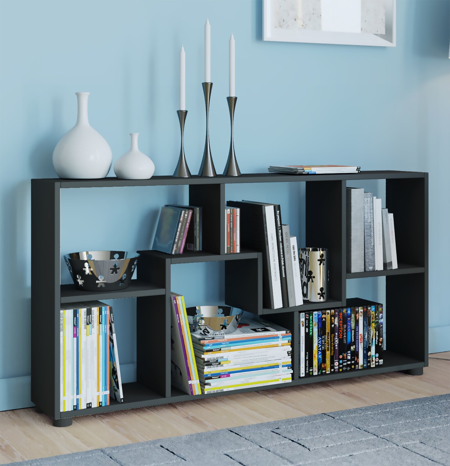 Modernes schwarzes Regal mit asymmetrischen Fächern, gefüllt mit Büchern und DVDs, dekoriert mit weißen Vasen und Kerzenhaltern; Frontansicht in leichter Perspektive von links vor hellblauer Wand.