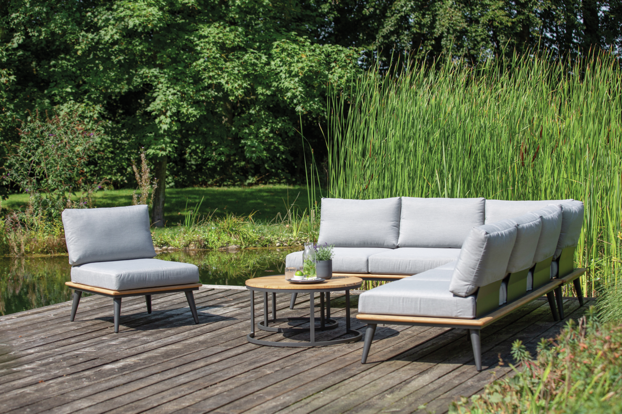 Moderne Outdoor-Lounge-Möbel auf einer Holzterrasse mit Blick auf einen Teich, bestehend aus einem Loungesessel, einer Loungebank und einem Loungetisch, umgeben von grüner Natur.