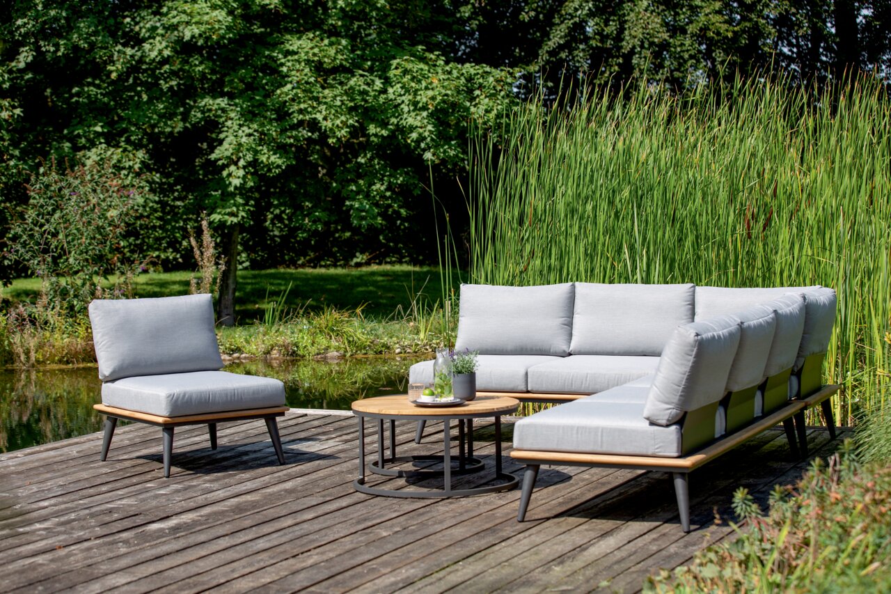 Moderne Outdoor-Lounge-Möbel auf einer Holzterrasse mit Blick auf einen Teich, bestehend aus einem Loungesessel, einer Loungebank und einem Loungetisch, umgeben von grüner Natur.