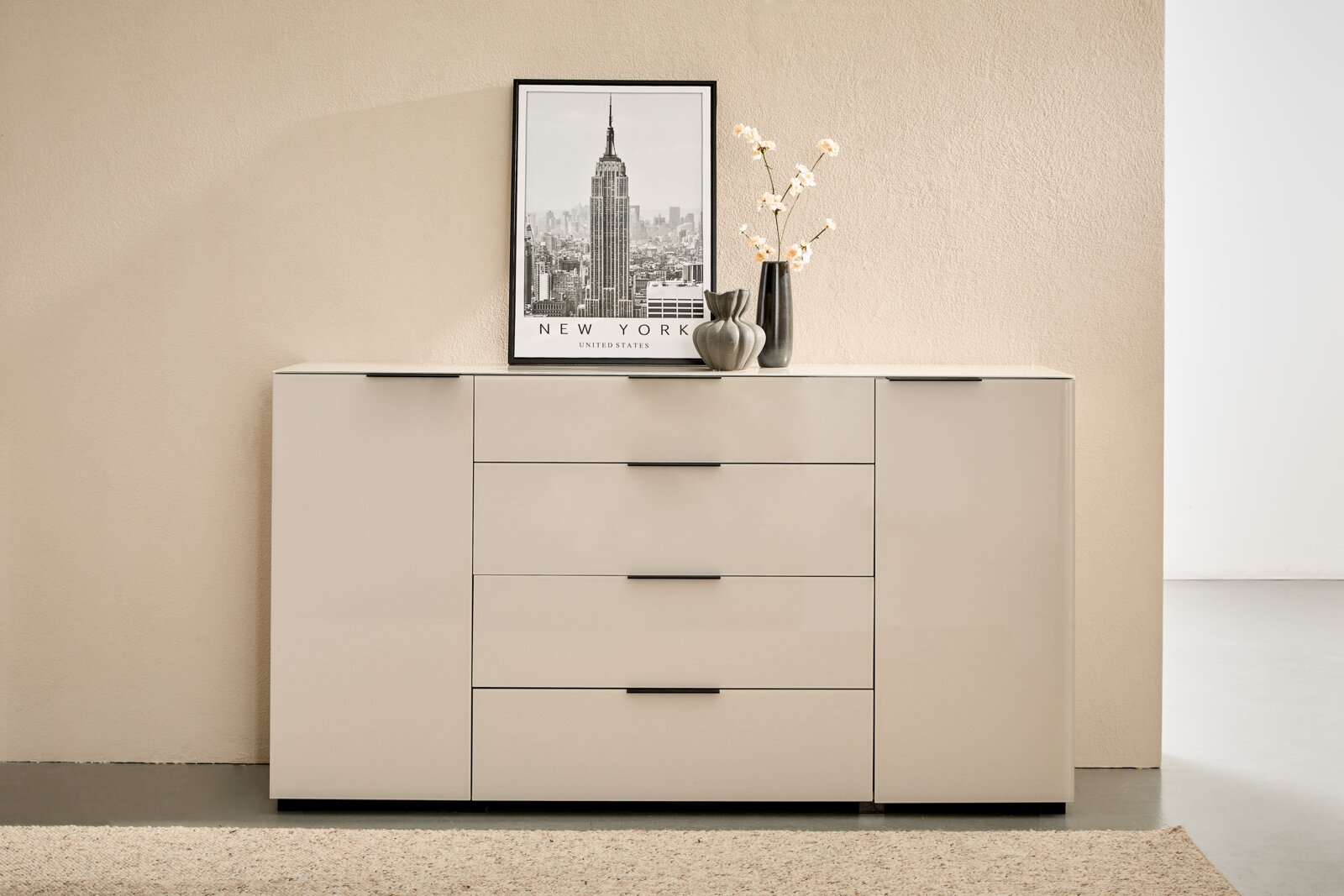 Novel Dressoir MALIN 2.0 Moderne witte hoogglans ladekast/dressoir met twee deuren en vier laden in het midden, zwarte handgrepen en decoratie (New York-poster, vazen met takken) op het oppervlak, van voren gefotografeerd vanuit een licht verhoogd perspectief