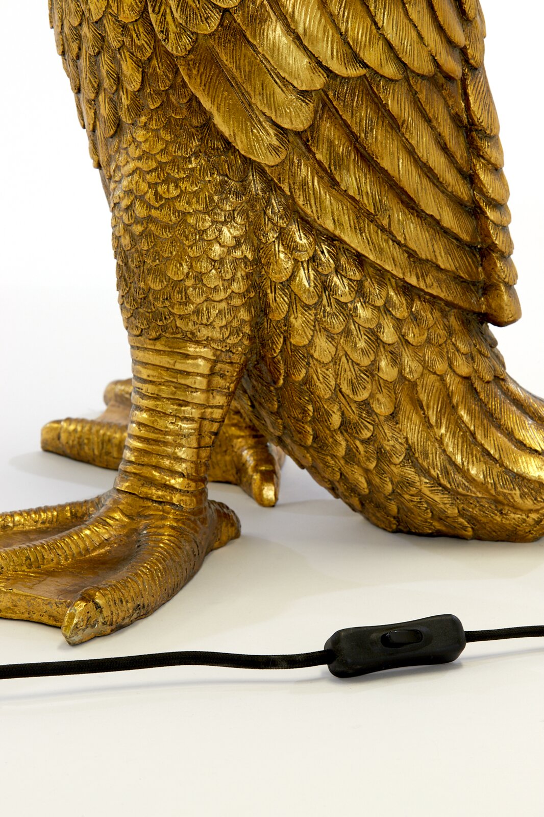 Close-up van een gouden tafellamp in de vorm van een vogel met gedetailleerde veren en een zwarte kabel met schakelaar op de voorgrond.