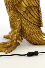 Close-up van een gouden tafellamp in de vorm van een vogel met gedetailleerde veren en een zwarte kabel met schakelaar op de voorgrond.