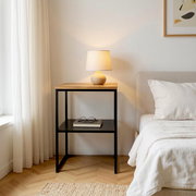 Moderner Nachttisch mit schwarzem Metallgestell und Holzplatte neben einem Bett, darauf eine leuchtende Tischlampe; frontale Perspektive im Schlafzimmer.