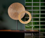 Zwei goldene Ornamente auf schwarzen Ständern, eines mit konzentrischen Kreisen und das andere als offener Ring, vor einer grünen Kachelwand, seitliche Perspektive.