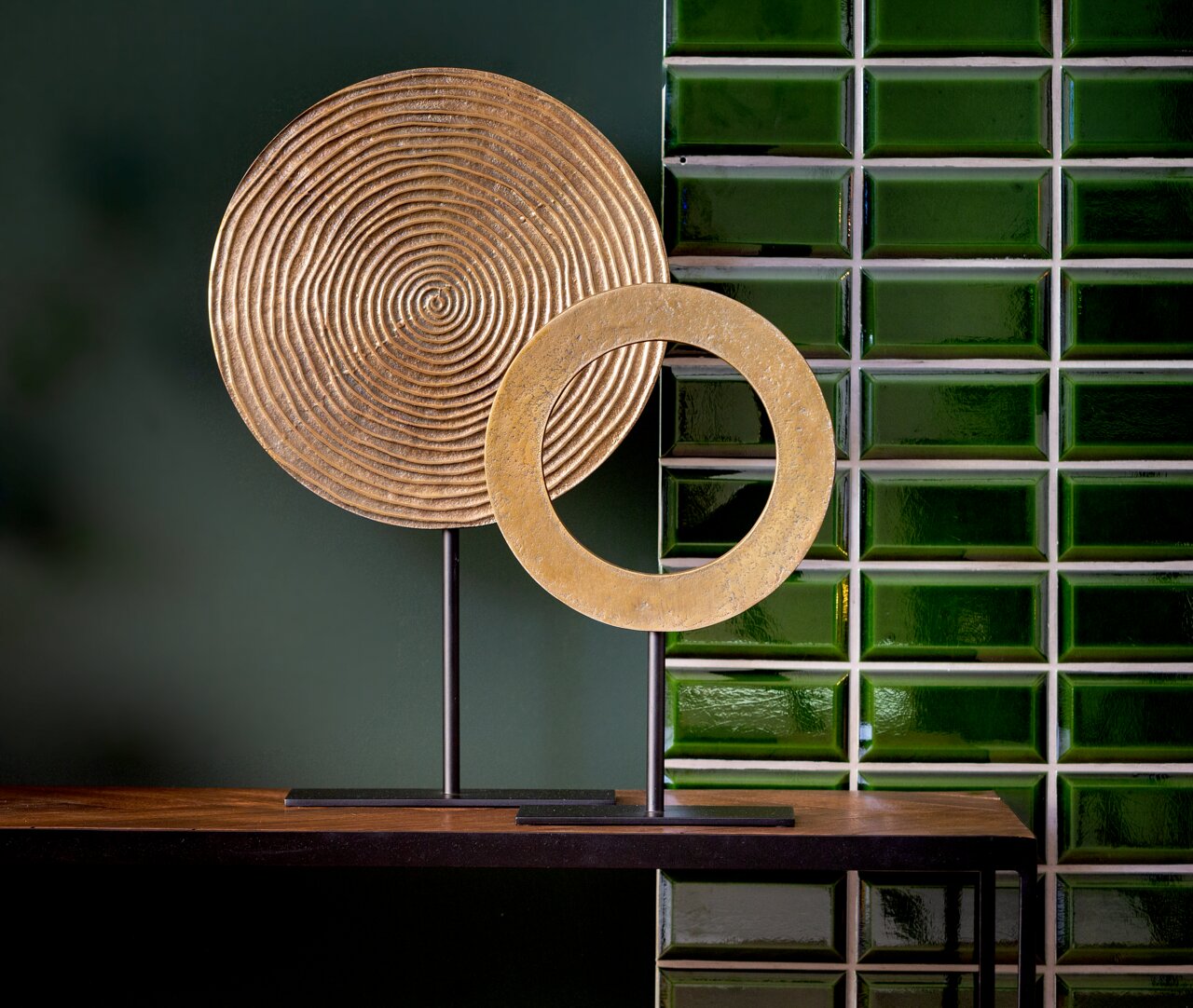 Zwei goldene Ornamente auf schwarzen Ständern, eines mit konzentrischen Kreisen und das andere als offener Ring, vor einer grünen Kachelwand, seitliche Perspektive.