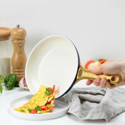 Eine Hand hält eine Pfanne mit goldfarbenem Griff, aus der ein Omelett mit Paprika und Kräutern auf einen Teller gleitet. Perspektive von der Seite.