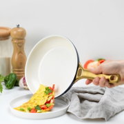 Een hand houdt een pan met een goudkleurig handvat vast, waaruit een omelet met paprika en kruiden op een bord glijdt. Zijaanzicht.