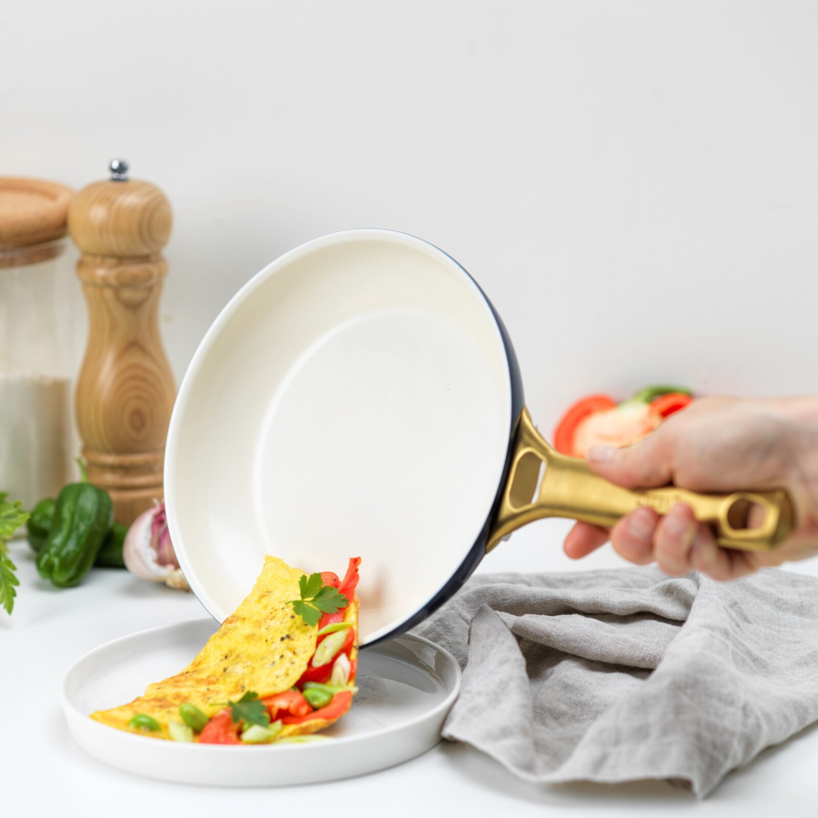 Een hand houdt een pan met een goudkleurig handvat vast, waaruit een omelet met paprika en kruiden op een bord glijdt. Zijaanzicht.