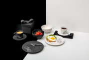 Schwarz-weißes Frühstücks-Set mit Tassen, Tellern und Schalen mit Cartoon-Motiv, angerichtet mit Kaffee, Erdbeeren und Toast mit Spiegelei auf zweifarbigem Hintergrund, Ansicht schräg von vorne/oben.