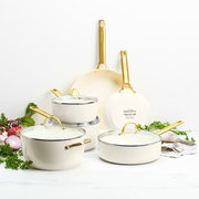Set aus cremefarbenen Töpfen und Pfannen mit goldenen Griffen, frontal fotografiert, umgeben von frischen Kräutern und Gemüse.