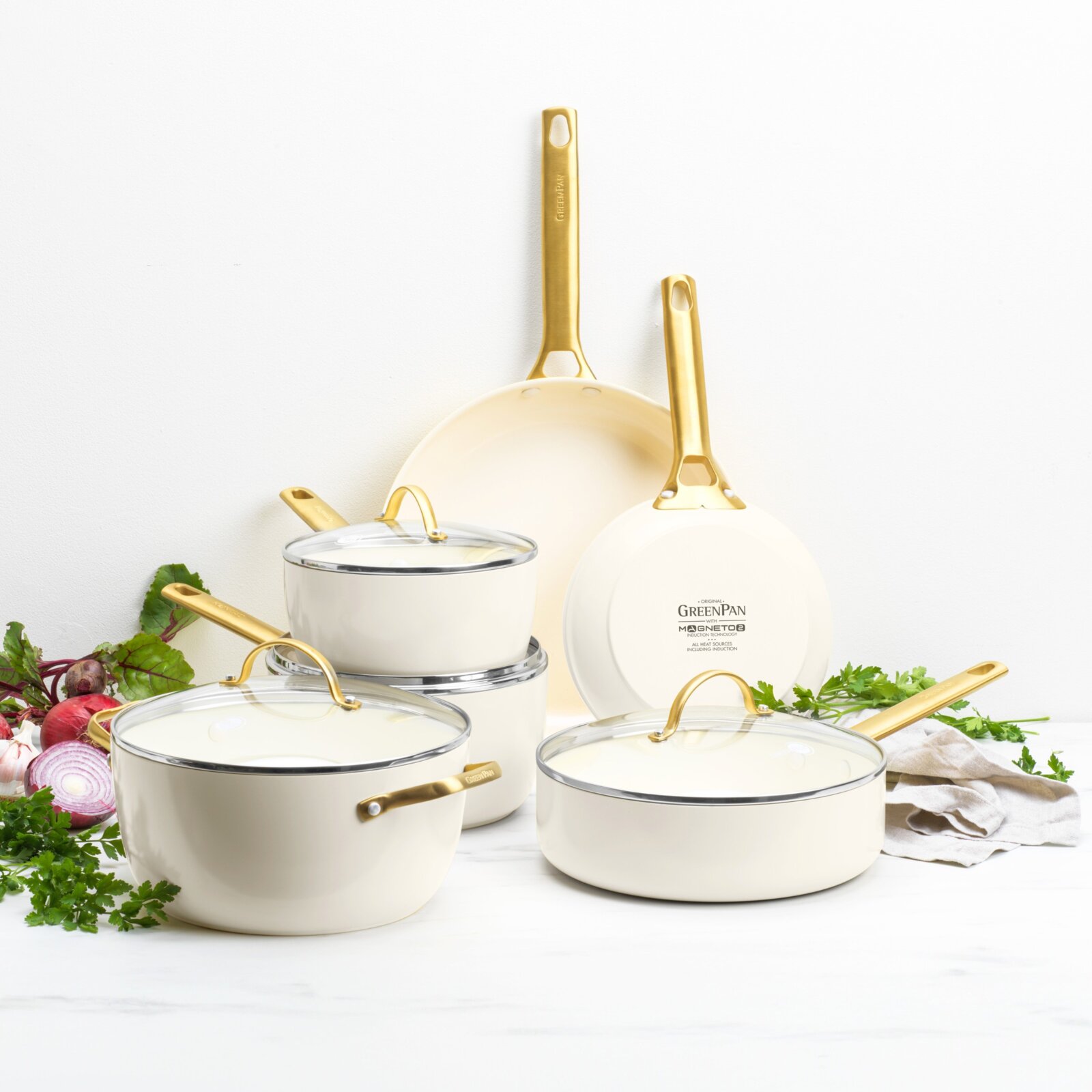 Set aus cremefarbenen Töpfen und Pfannen mit goldenen Griffen, frontal fotografiert, umgeben von frischen Kräutern und Gemüse.