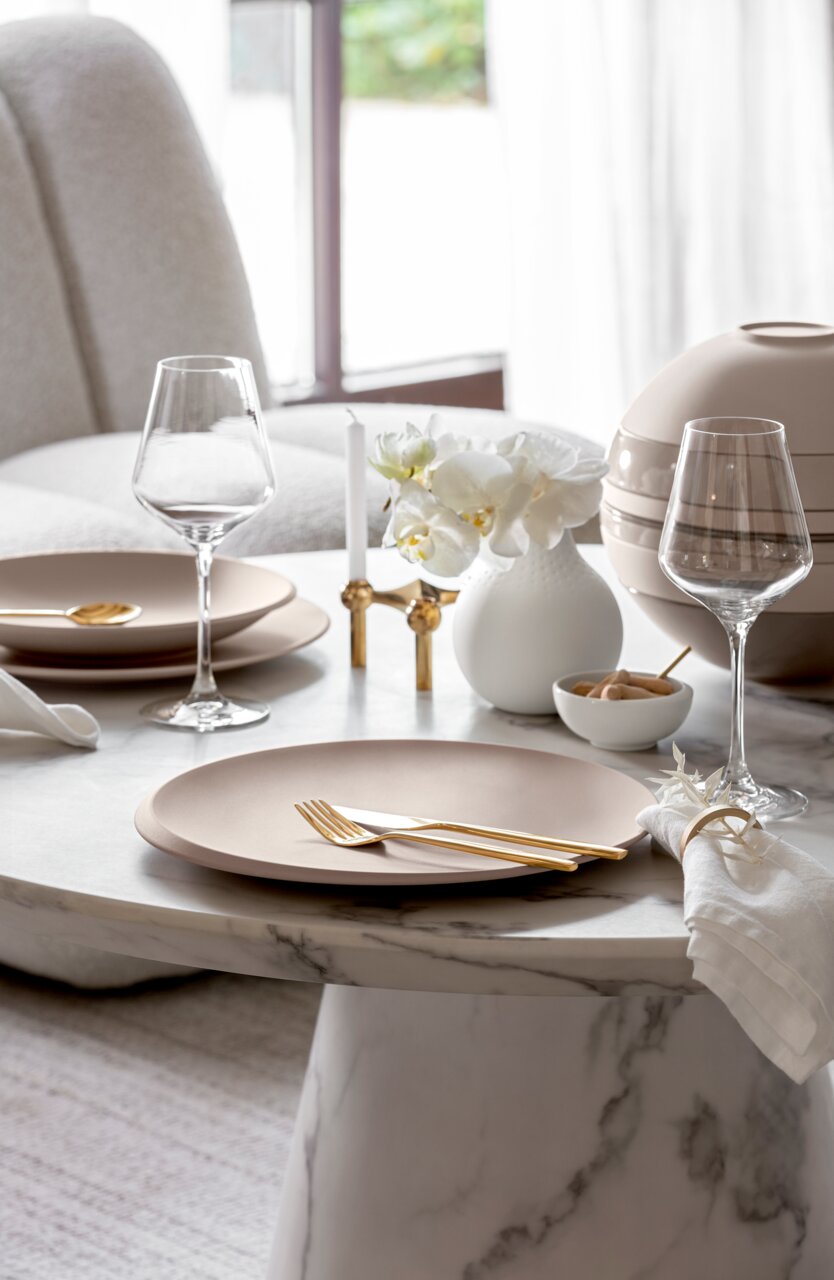 Elegante Tischdekoration mit beiger Schale NewMoon 750ml, goldfarbenem Besteck und Weingläsern auf einem Marmortisch, seitliche Perspektive.