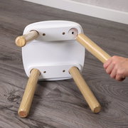 Unteransicht eines kleinen weißen Hockers mit abnehmbaren Holzbeinen; eine Hand schraubt ein Bein in die Halterung, auf grauem Holzboden.