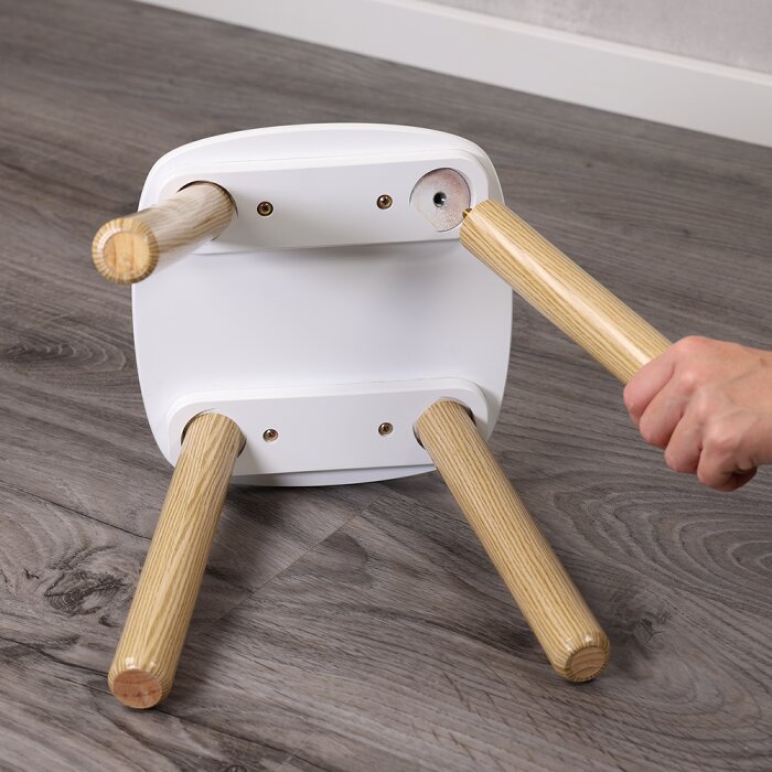 Unteransicht eines kleinen weißen Hockers mit abnehmbaren Holzbeinen; eine Hand schraubt ein Bein in die Halterung, auf grauem Holzboden.