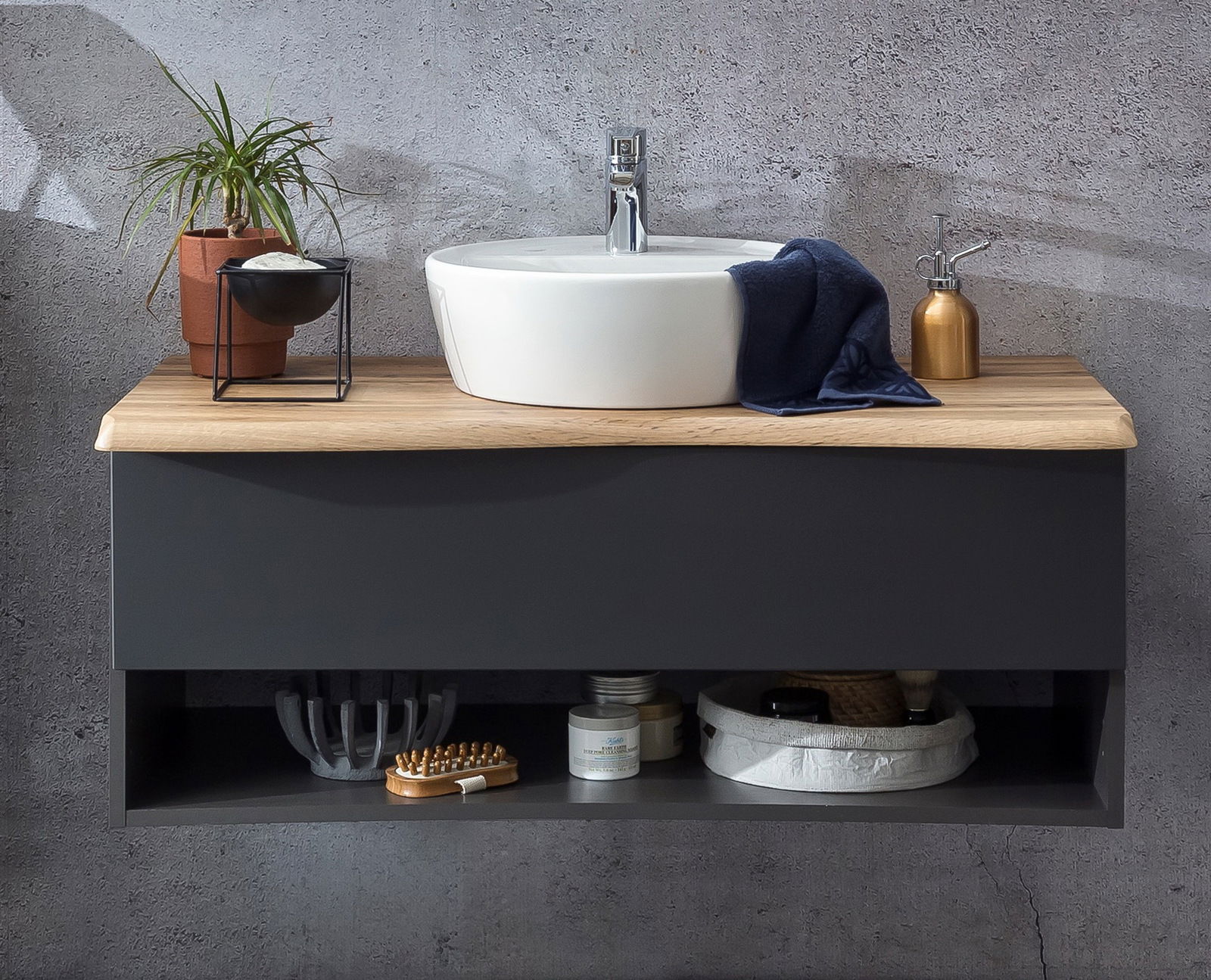 Moderner Waschtisch mit rundem weißen Waschbecken auf einer Holzplatte, dekoriert mit einer Pflanze, Seifenspender und Handtuch, frontal betrachtet.