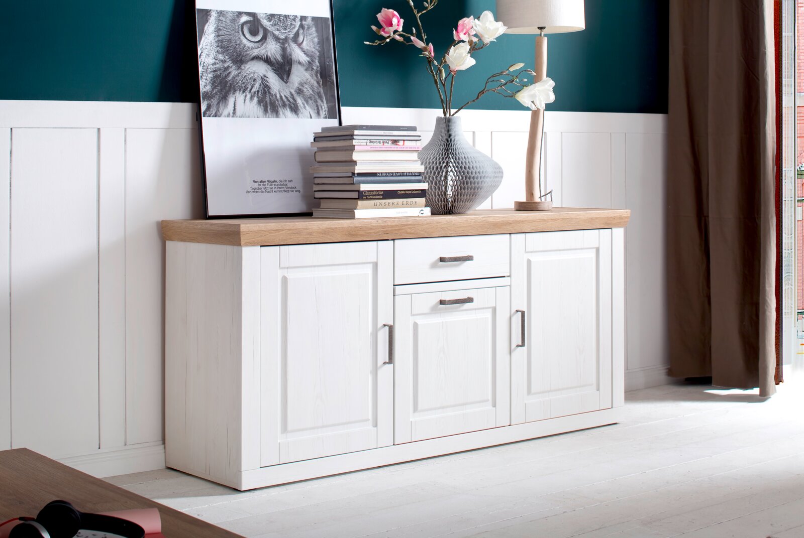 Weißes Sideboard mit Holzplatte und Metallgriffen, dekoriert mit Büchern, Bild, Vase und Tischlampe; Perspektive: frontale Ansicht in leichter Schrägansicht von links.