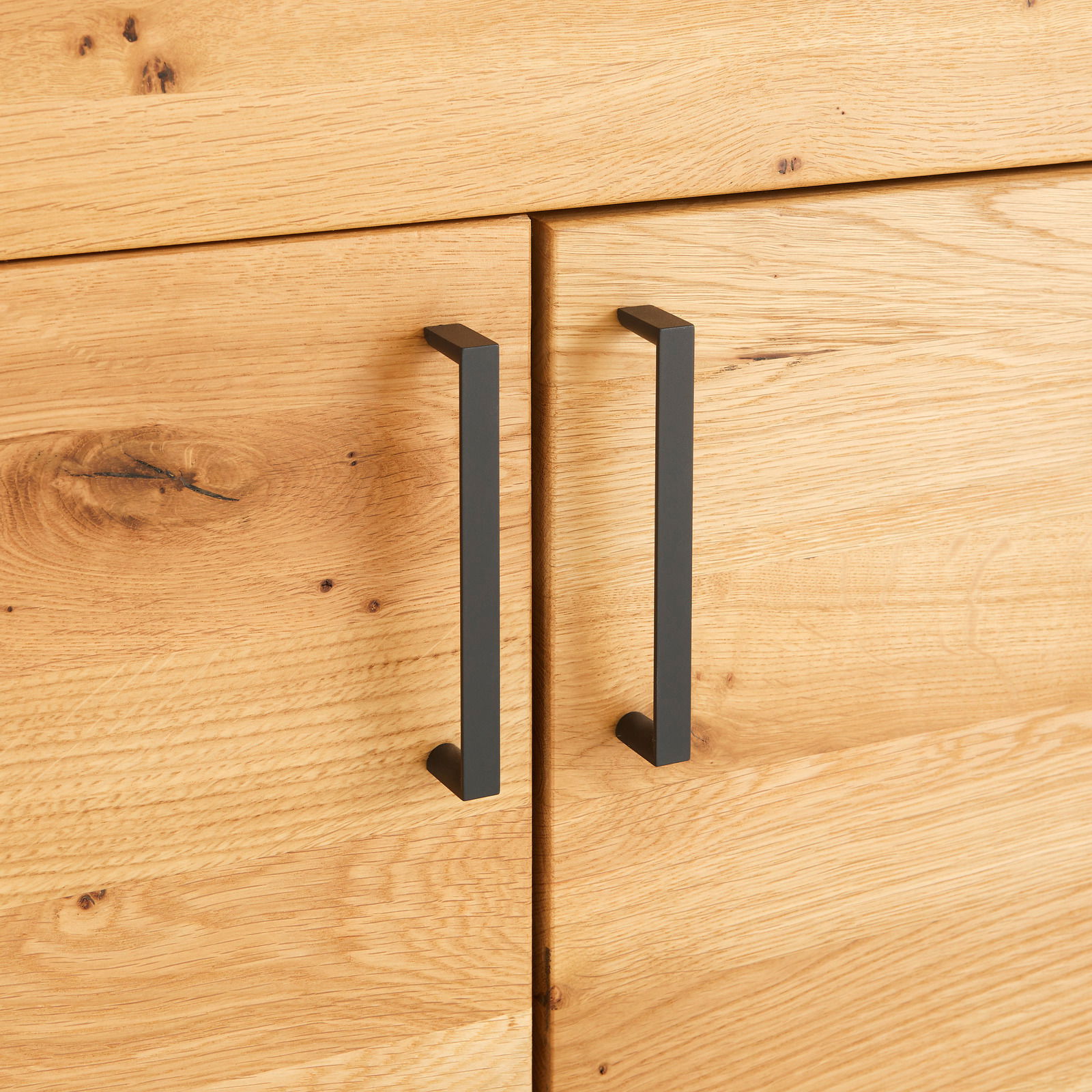 Nahaufnahme von zwei schwarzen, modernen Griffen an einer Holzkommode aus hellem Holz mit sichtbarer Maserung, seitliche Perspektive.