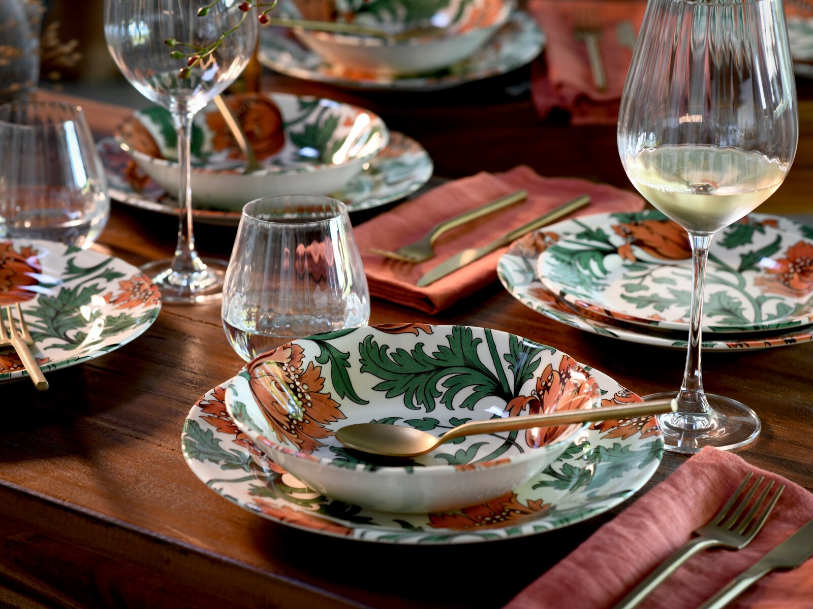 Gedeckter Holztisch mit floral gemustertem Porzellan-Geschirr, goldfarbenem Besteck, Gläsern und Weißweinglas, schräg von oben aus der Nähe fotografiert.