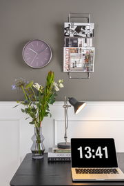 Wandmontiertes Zeitungsregal mit mehreren Zeitschriften, daneben eine Wanduhr. Perspektive: Frontalansicht.