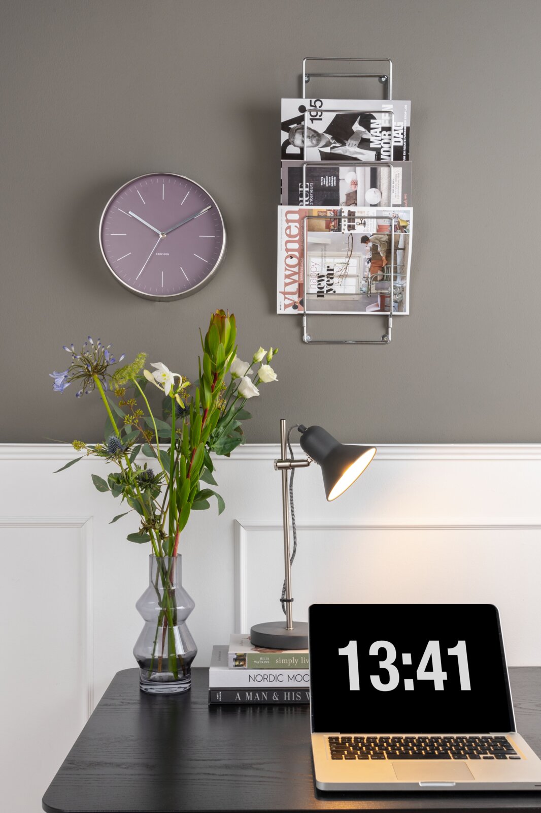 Een aan de muur bevestigd krantenrek met verschillende tijdschriften, met daarnaast een wandklok. Perspectief: vooraanzicht.