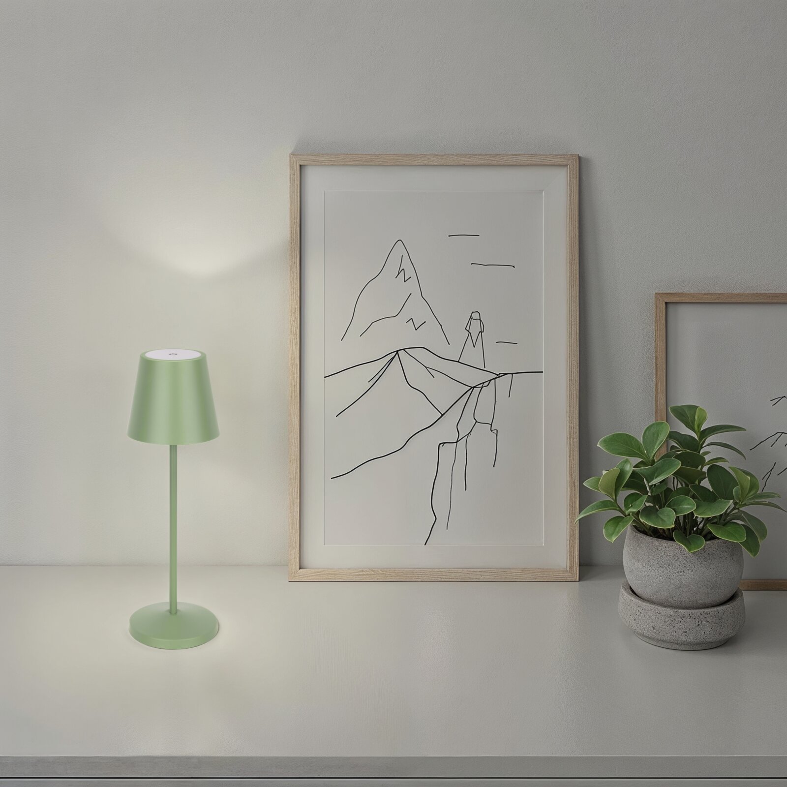Minimalistisches gerahmtes Poster mit Linienzeichnung einer Berglandschaft, frontal aus leichter Seitenperspektive fotografiert; daneben eine grüne Tischlampe und eine Topfpflanze auf einem Regal.