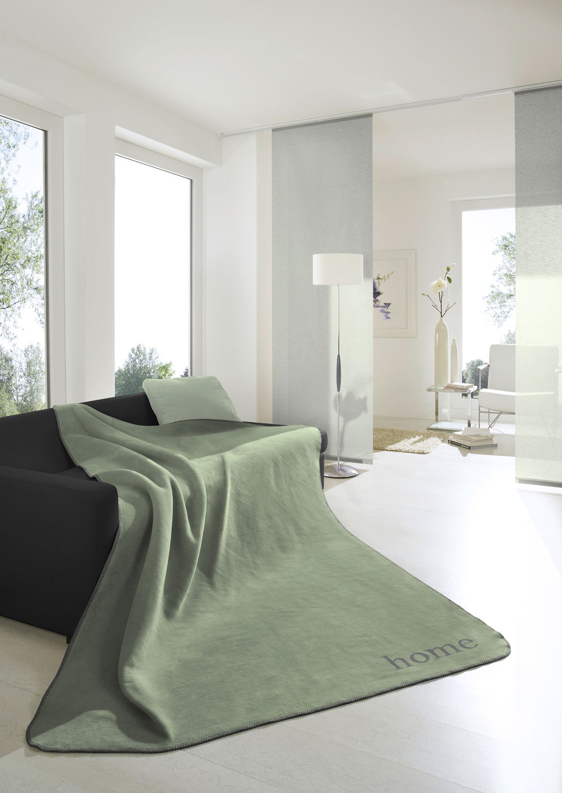 Grüne Wohndecke 150x200cm auf einem schwarzen Sofa in einem hellen Wohnzimmer, seitliche Perspektive