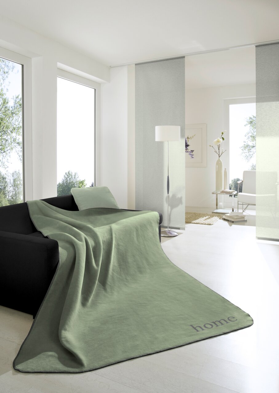 Grüne Wohndecke 150x200cm auf einem schwarzen Sofa in einem hellen Wohnzimmer, seitliche Perspektive